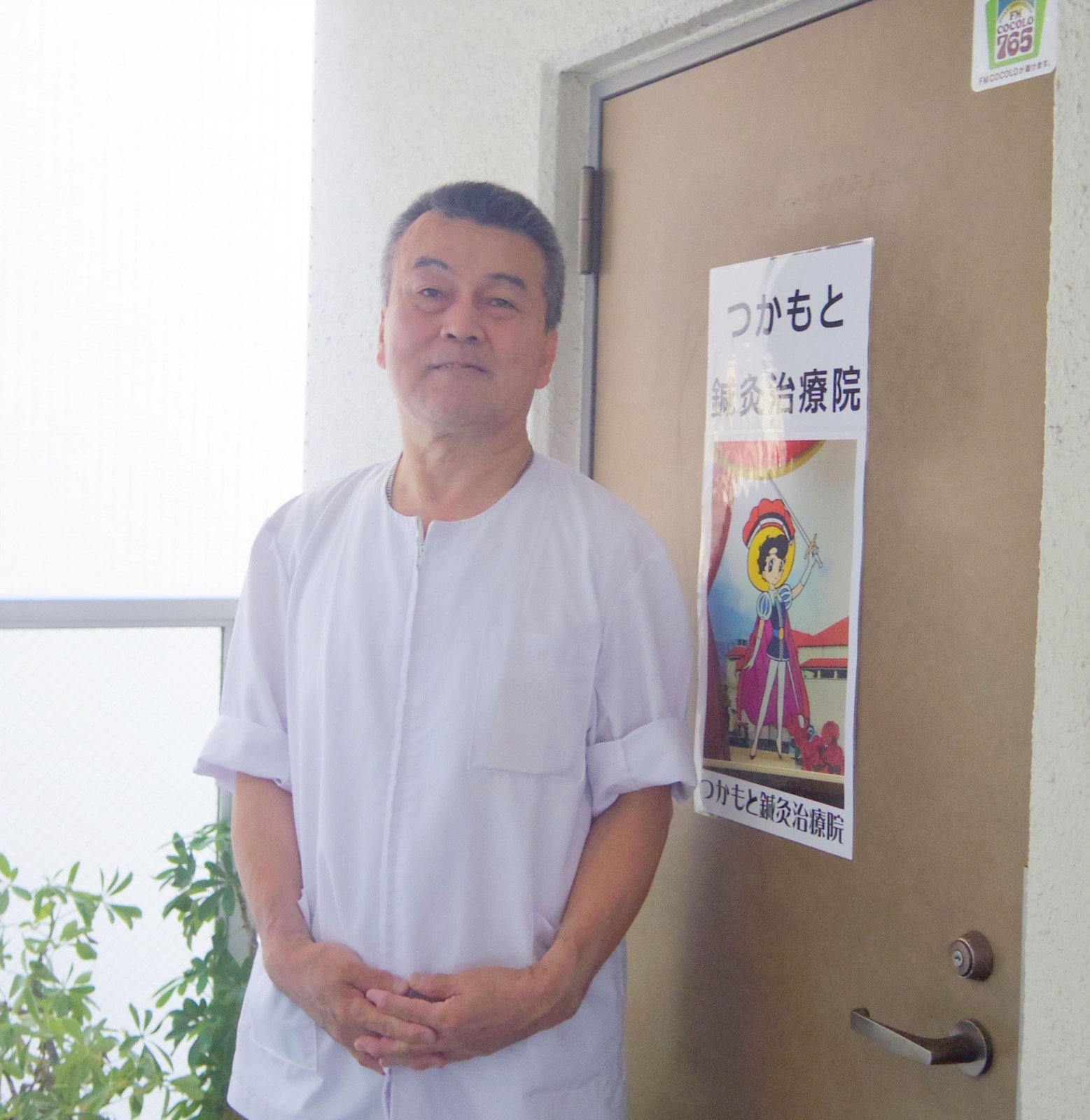 つかもと鍼灸治療院の表示が貼られた605号室の入口ドアと、その前に立つ院長